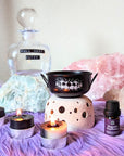Black Mystic cauldron with scented candles and protection lotion on purple rug and ritual crystals in the background. Available at Goddess Provisions