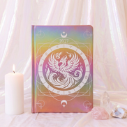 A hardcover journal with a 2027 calendar cover design, featuring a white phoenix and zodiac wheel motif over an iridescent pastel gradient background. The journal is styled on a draped white fabric surface next to a lit white candle and crystals.