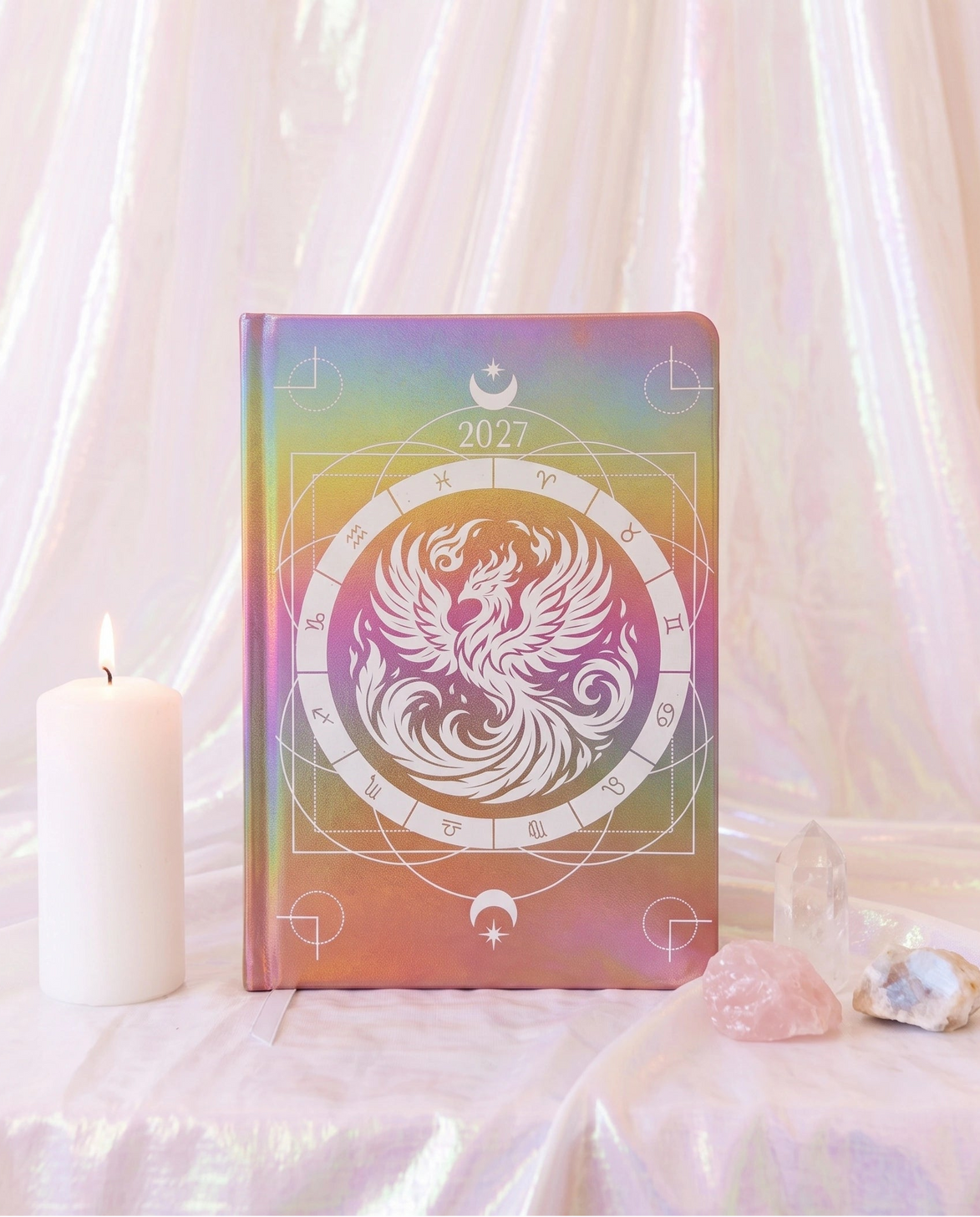 A hardcover journal with a 2027 calendar cover design, featuring a white phoenix and zodiac wheel motif over an iridescent pastel gradient background. The journal is styled on a draped white fabric surface next to a lit white candle and crystals.