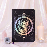 A black hardcover 2027 journal featuring a central iridescent phoenix design with a zodiac wheel, styled on draped white fabric with a pink crystal, a large white candle, and a tea light.