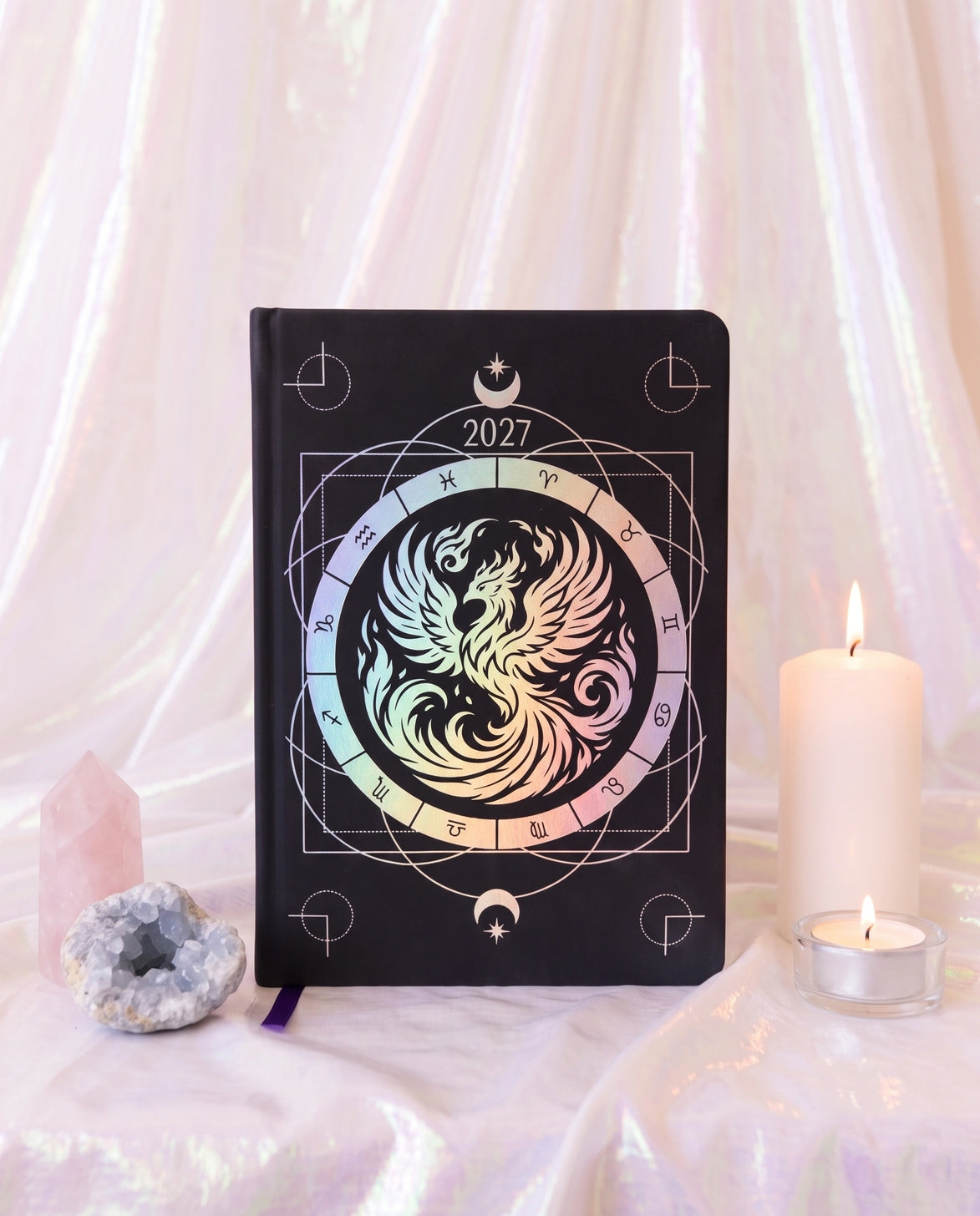 A black hardcover 2027 journal featuring a central iridescent phoenix design with a zodiac wheel, styled on draped white fabric with a pink crystal, a large white candle, and a tea light.
