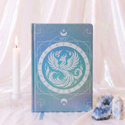A hardcover 2027 zodiac planner or journal with a blue and teal phoenix design, styled on a draped white fabric surface with a lit candle and crystals.