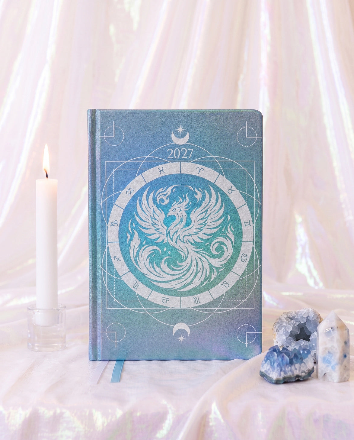 A hardcover 2027 zodiac planner or journal with a blue and teal phoenix design, styled on a draped white fabric surface with a lit candle and crystals.