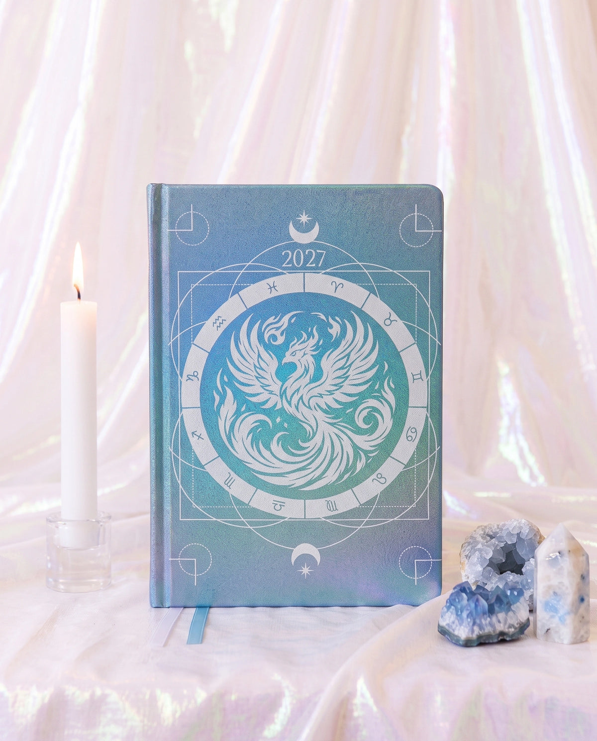 A hardcover 2027 zodiac planner or journal with a blue and teal phoenix design, styled on a draped white fabric surface with a lit candle and crystals.