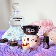 Black Mystic cauldron with scented candles and protection lotion on purple rug and ritual crystals in the background. Available at Goddess Provisions