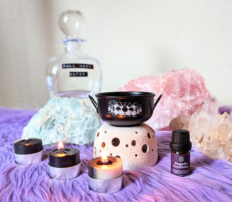 Black Mystic cauldron with scented candles and protection lotion on purple rug and ritual crystals in the background. Available at Goddess Provisions