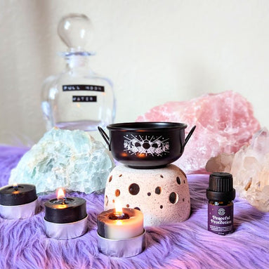 Black Mystic cauldron with scented candles and protection lotion on purple rug and ritual crystals in the background. Available at Goddess Provisions