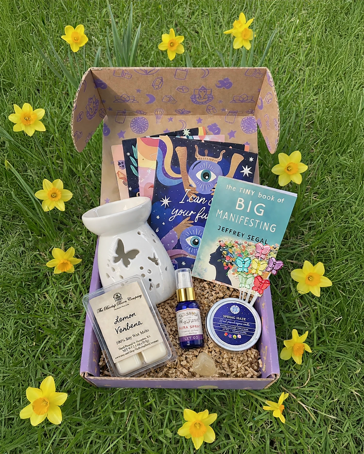 Box of self-care items including a diffuser, books, and candles on grass with daffodils.