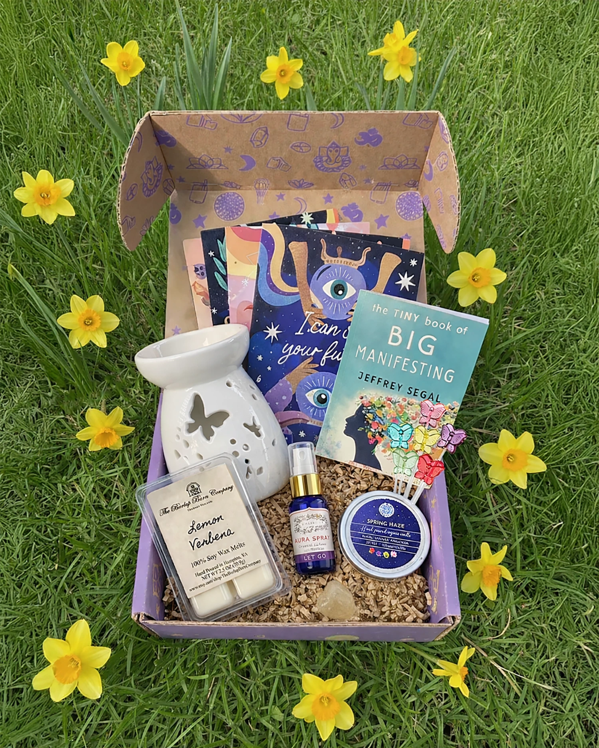 Box of self-care items including a diffuser, books, and candles on grass with daffodils.
