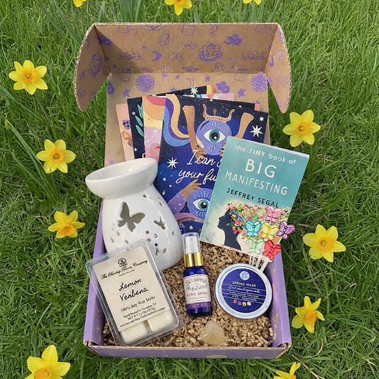 Box of self-care items including a diffuser, books, and candles on grass with daffodils.