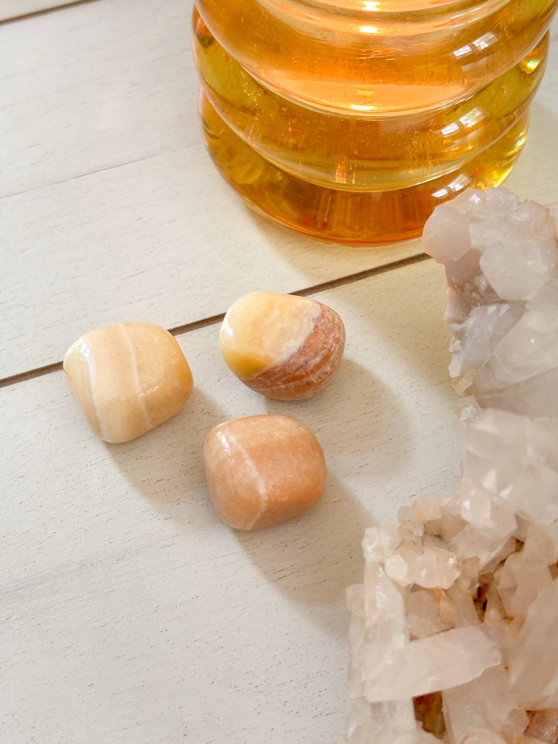 Three tumbled stones and a crystal cluster on a light surface with a glass container of honey in the background.