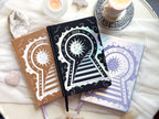 Three decorative 2026 Moon Planners / notebooks with 2026 design on a white surface with candles and crystals. 