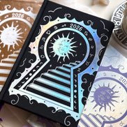 Stunning black planner decorative 2026 Moon Planners / notebooks with 2026 design on a white surface with candles and crystals. Goddess Provisions Planner - Exclusive. With champaign 2026 planner and Silver 2026 planner in the background. 