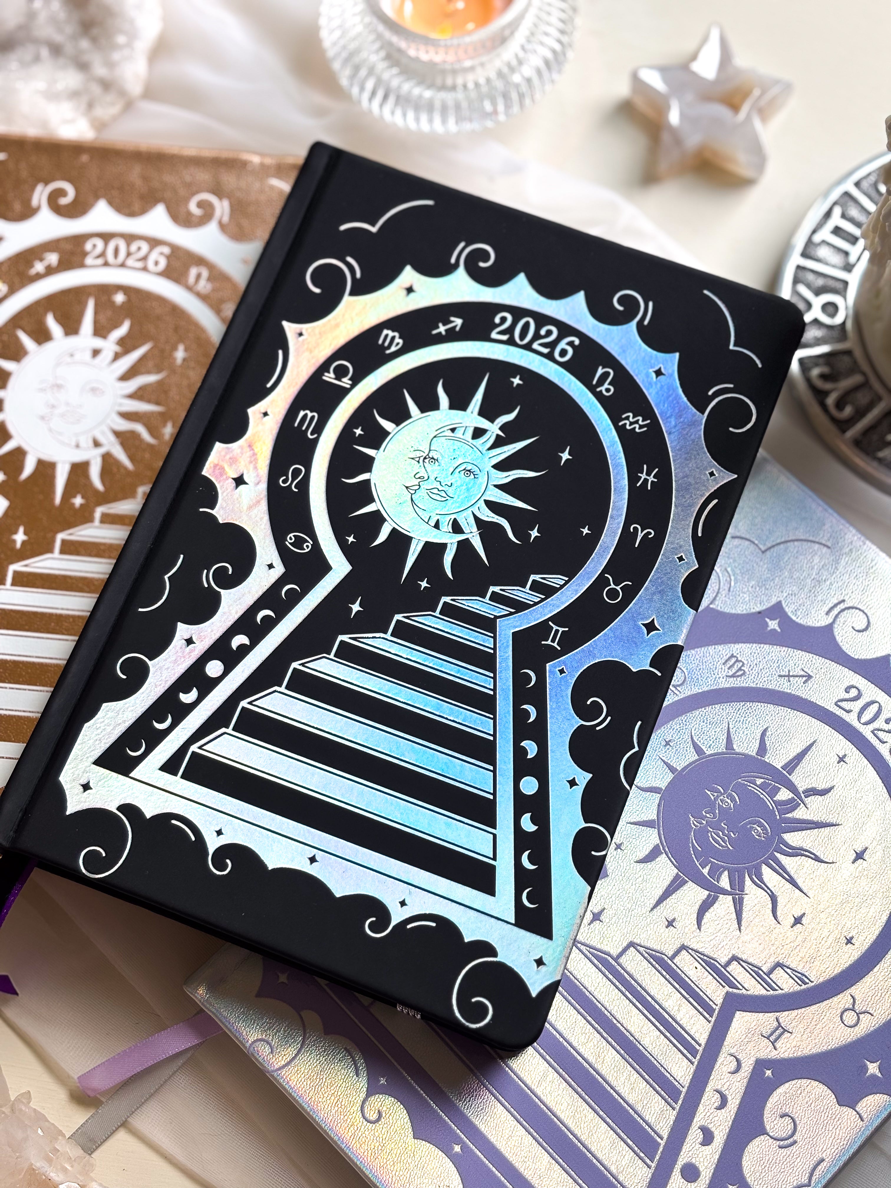 Stunning black planner decorative 2026 Moon Planners / notebooks with 2026 design on a white surface with candles and crystals. Goddess Provisions Planner - Exclusive. With champaign 2026 planner and Silver 2026 planner in the background. 