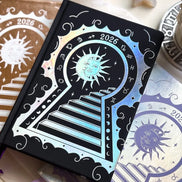 Stunning black planner decorative 2026 Moon Planners / notebooks with 2026 design on a white surface with candles and crystals. Goddess Provisions Planner - Exclusive. With champaign 2026 planner and Silver 2026 planner in the background. 