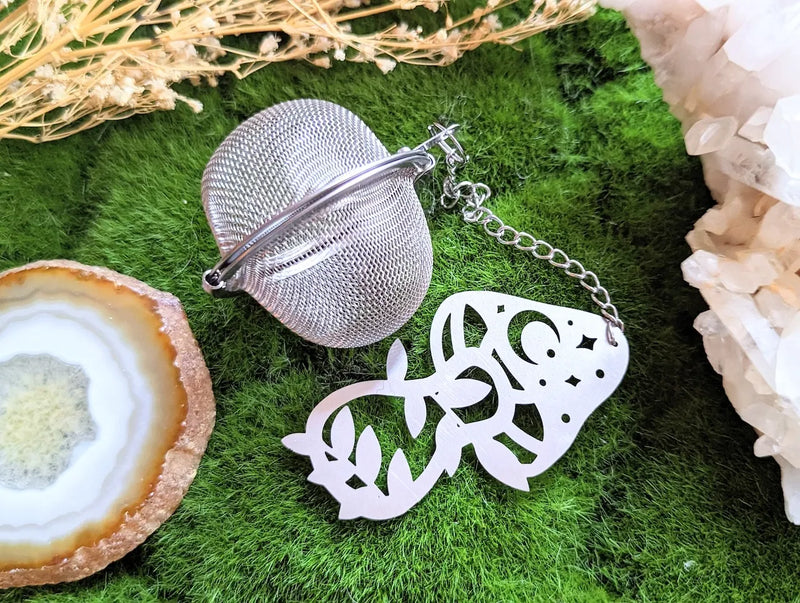 Herbal Alchemy tea infuser with stainless steel mesh ball and whimsical mushroom charm beside loose leaf tea.