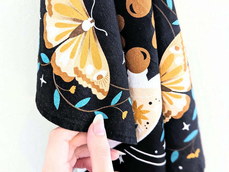 Black cotton tea towel featuring mystical moon and star designs styled beside candles and plants.