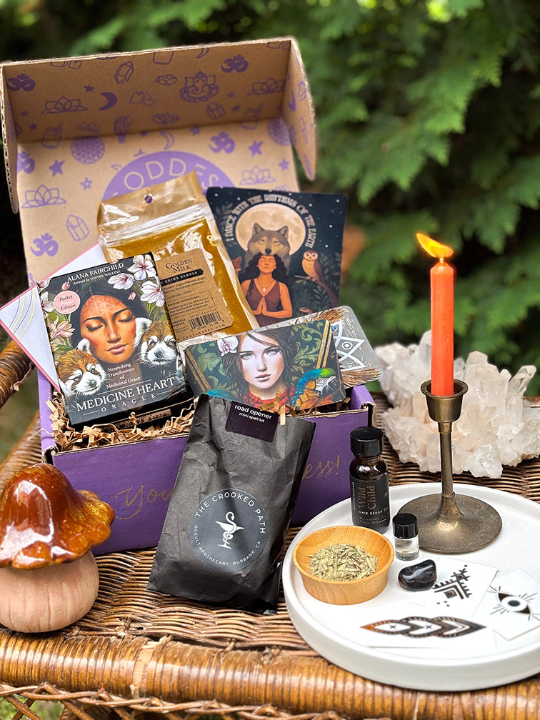 A Goddess Provisions box with oracle cards, a candle, crystals and sage on a wicker table