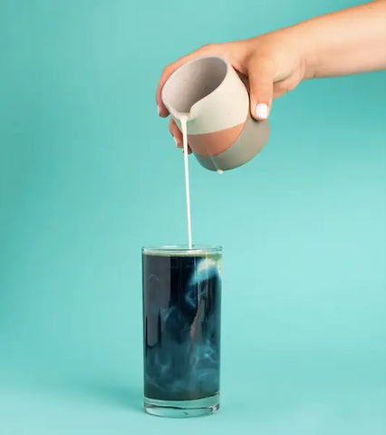 Hand pouring white liquid from a pitcher into a glass of dark blue liquid for Herbal Allies for Energy article