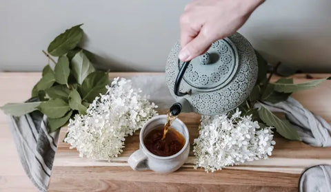 Ornate teal teapot pouring dark herbal tea into a small mug for Root Chakra support