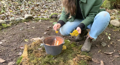 Person in green jacket and blue jeans adding yellow plant material to a pot with fire for plant alchemy
