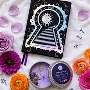 Black 2026 moon planners or 2026 Lunar planners with a Purple spring Haze candle with herbs inside and flowers on a light cream silk surface