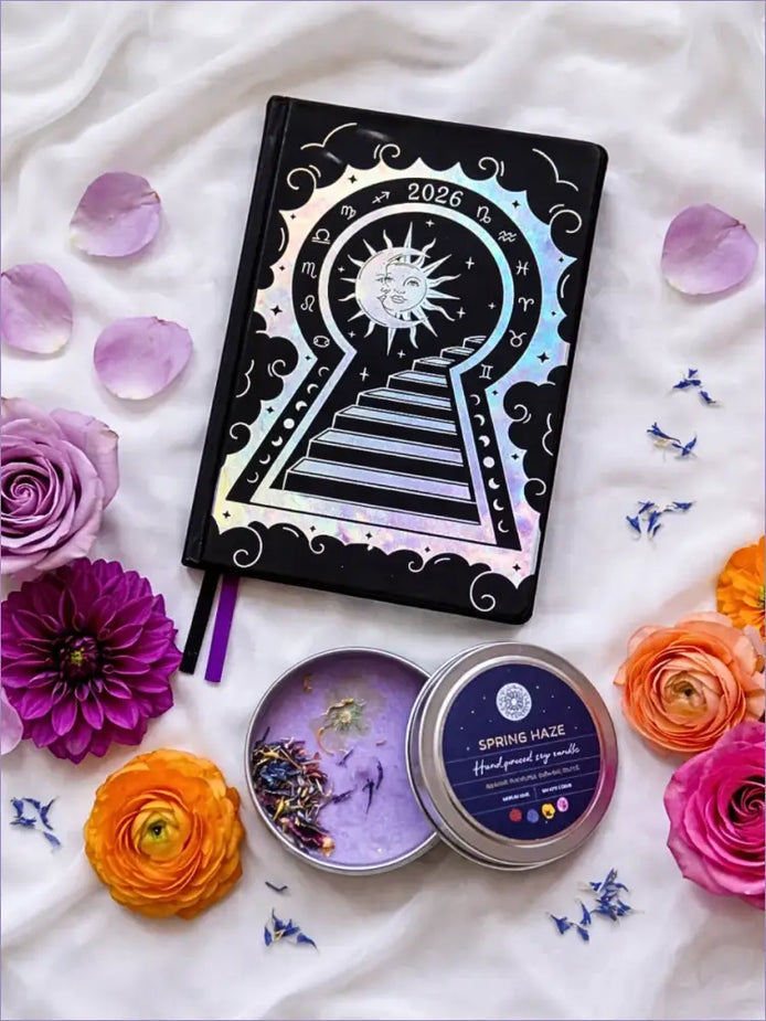 Black 2026 moon planners or 2026 Lunar planners with a Purple spring Haze candle with herbs inside and flowers on a light cream silk surface