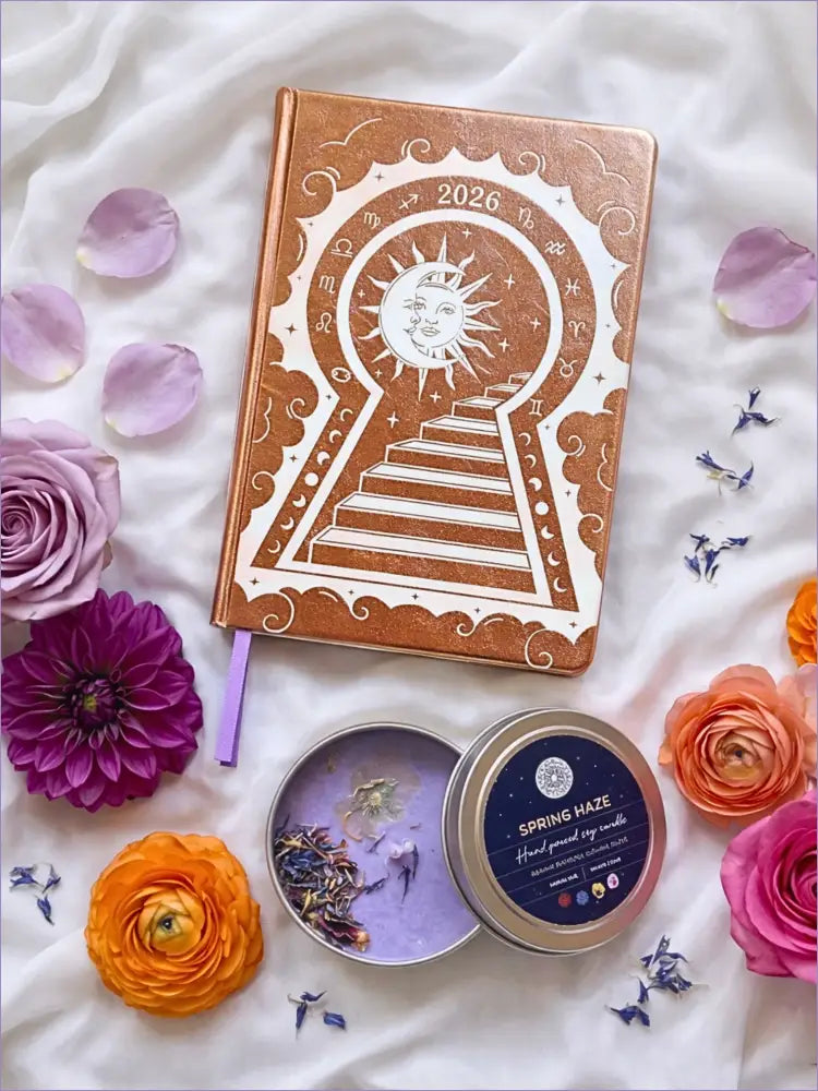 Gold 2026 moon planners or 2026 Lunar planners with a spring Haze candle with herbs inside and flowers on a light cream silk surface