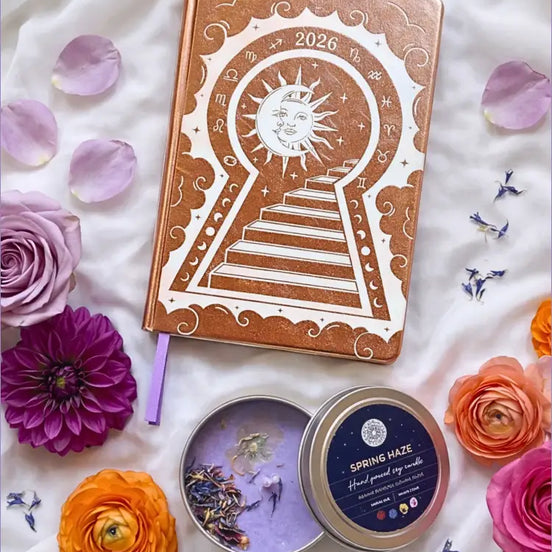 Gold 2026 moon planners or 2026 Lunar planners with a spring Haze candle with herbs inside and flowers on a light cream silk surface