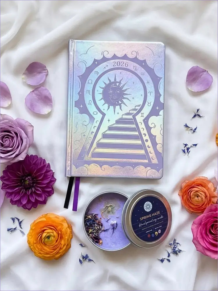 Three Silver 2026 moon planners or 2026 Lunar planners with a purple spring Haze candle with herbs inside and flowers on a light cream silk surface