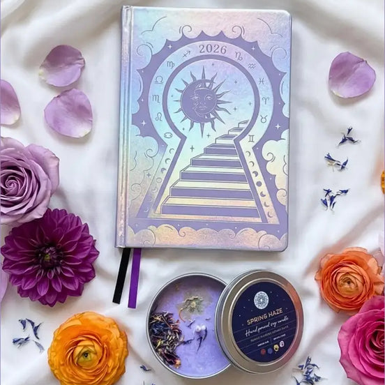Three Silver 2026 moon planners or 2026 Lunar planners with a purple spring Haze candle with herbs inside and flowers on a light cream silk surface