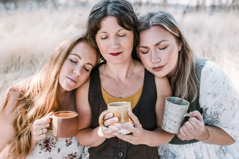 Three women with eyes closed holding ceramic mugs in a calming herbal wellness setting