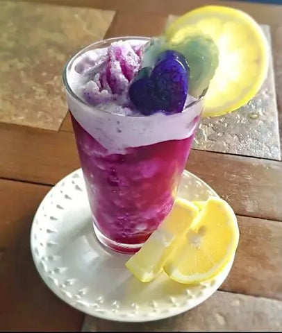 Vibrant purple beverage with frothy white top, edible flowers, lemon slices in clear glass