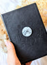 Mystical Musings Divination Diary with black vegan leather cover, featuring embossed clear quartz crystal and moon phases design. A celestial journal for dreams and manifestations. Available at Goddess Provisions.