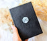 Mystical Musings Divination Diary with black vegan leather cover, featuring embossed clear quartz crystal and moon phases design. A celestial journal for dreams and manifestations. Available at Goddess Provisions.
